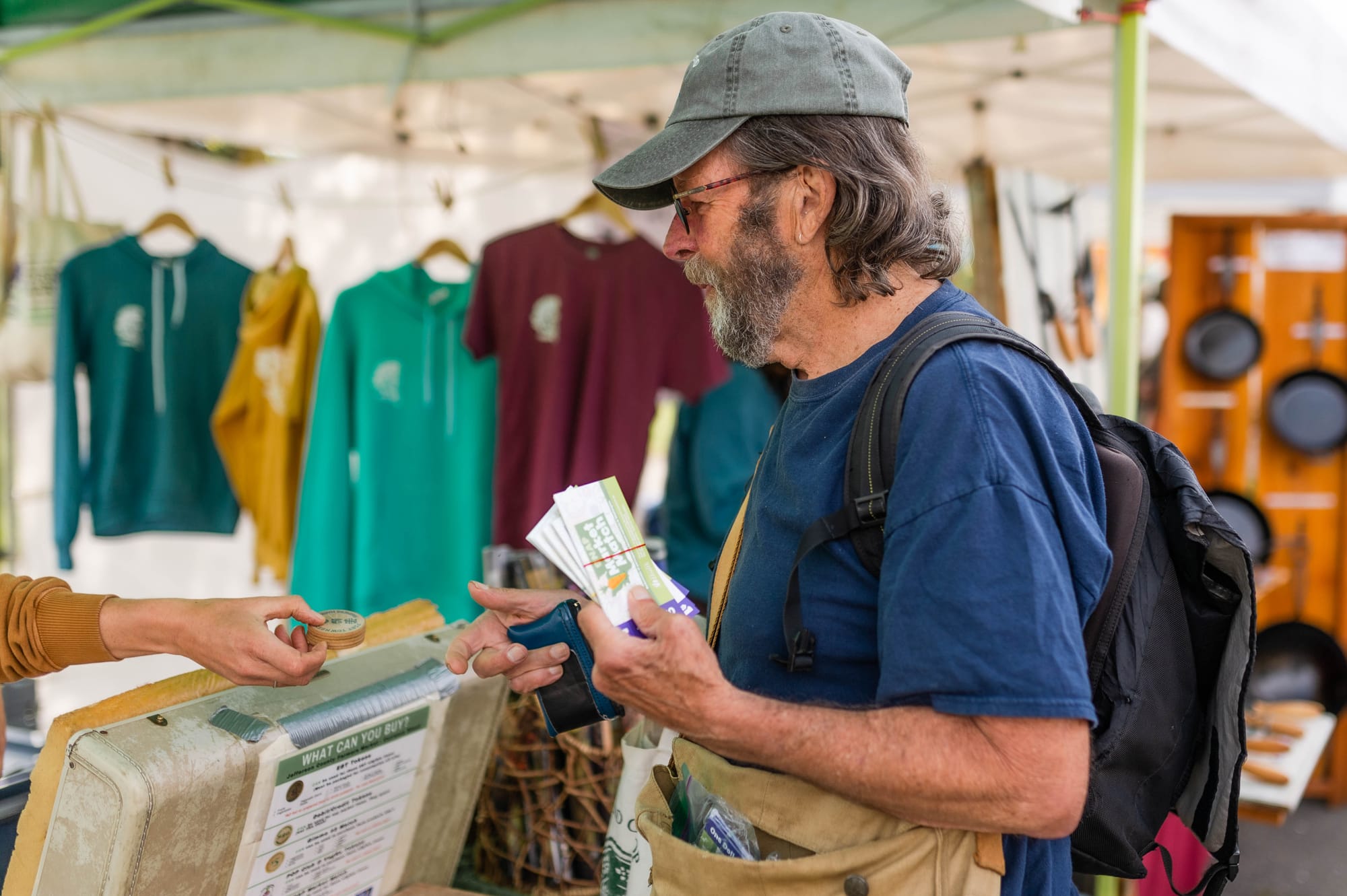 A man in a blue shirt and gray hat holding Farmer's Market currency in the midst of a transaction. 