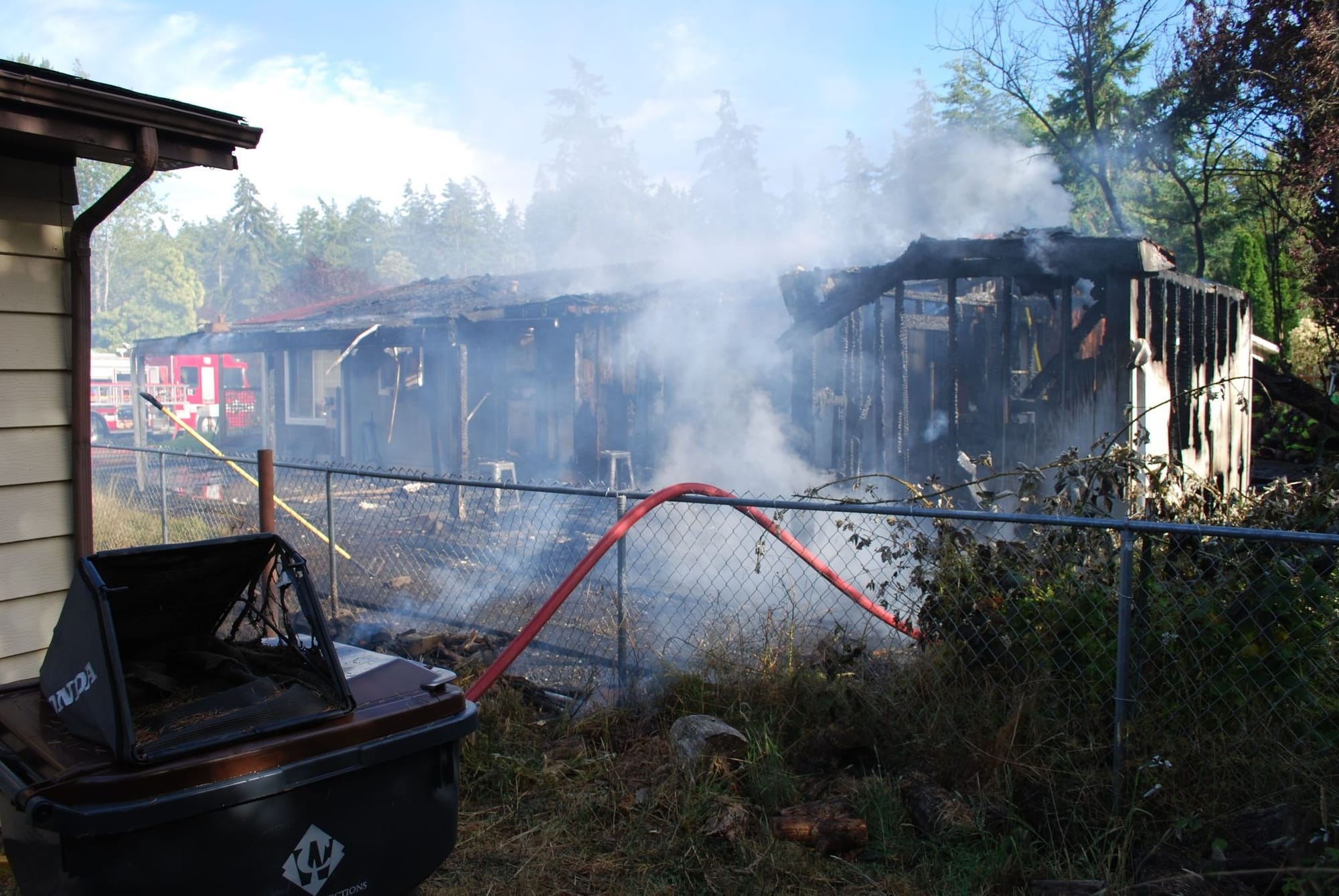 A very burnt and smoldering house behind a chainlink fence with a firehose draped over it.