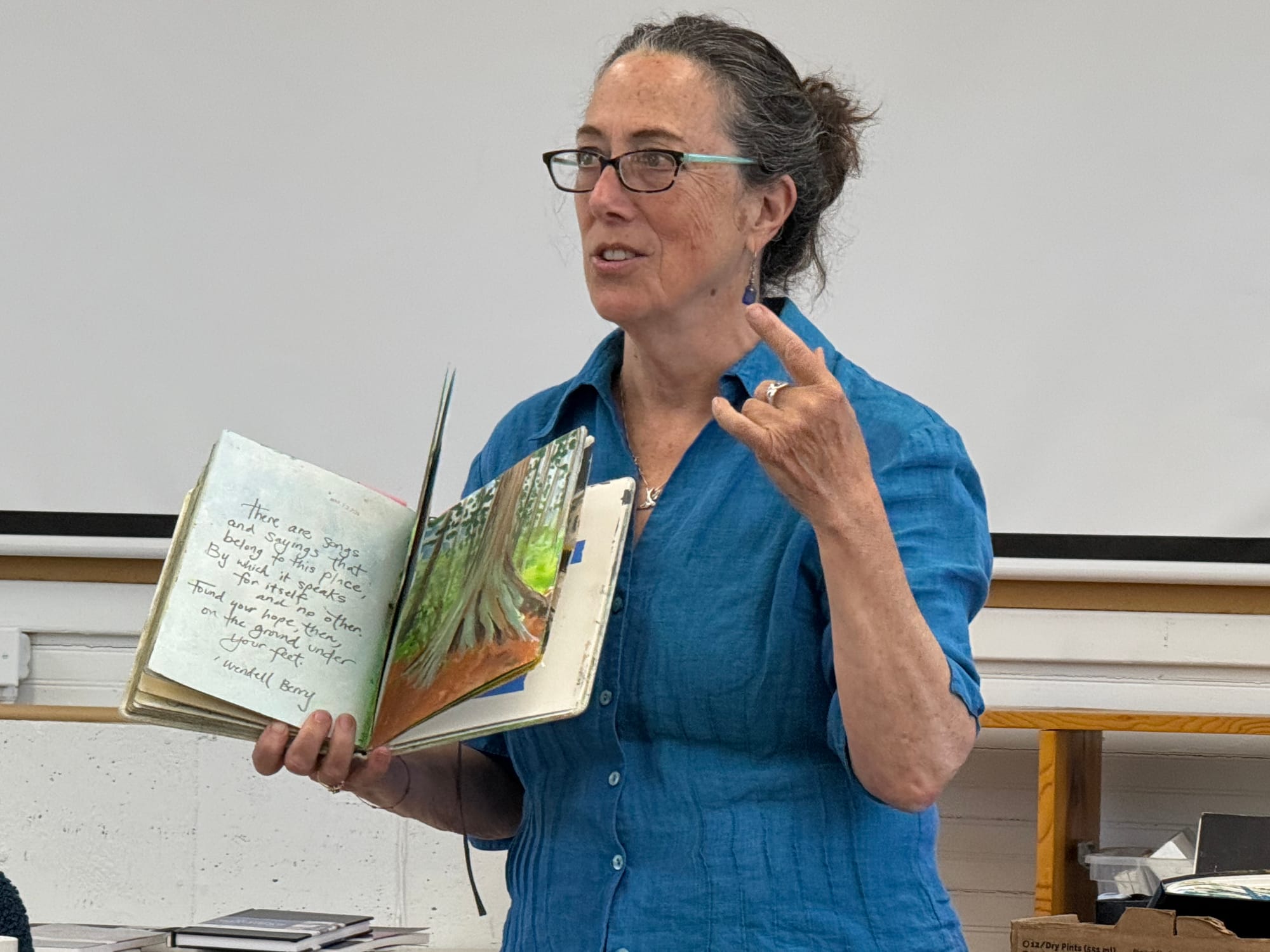 Teaching artist, author and painter Meg Kaczyk holding an illustrated book and gesturing with her hand. 