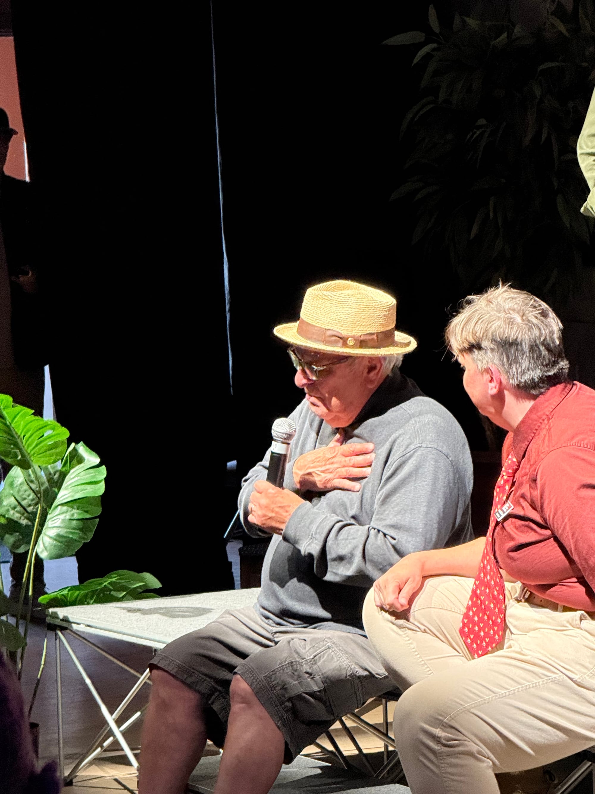 Conrad Lozano, bassist and founding member of Los Lobos, left, speaking with his hand to his heart into a microphone with Festival executive director Danielle McClelland listening beside him at the front of the American Legion stage. 
