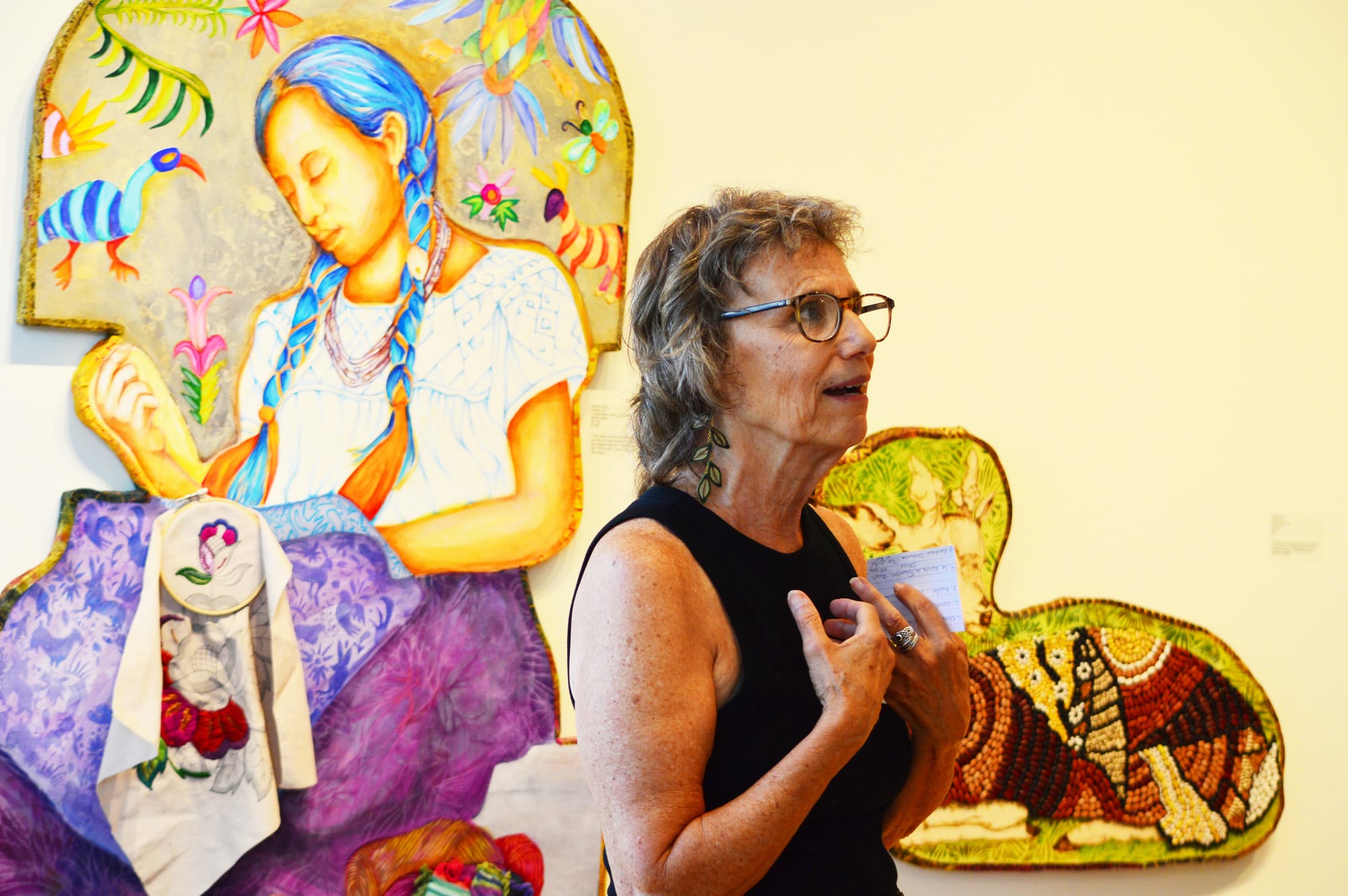 Carolyn King discusses “La Bordadera Otomi” standing in front of a painting of a woman sewing.