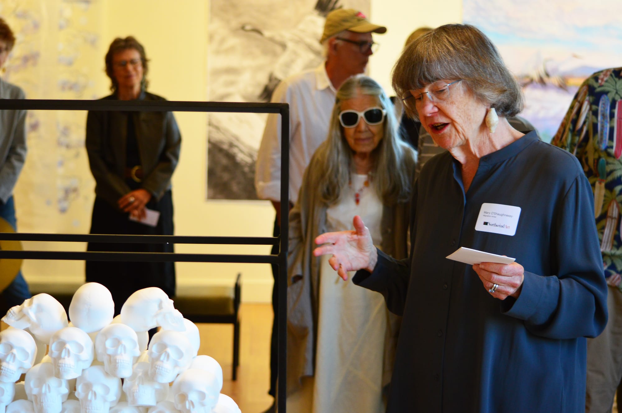 Artist Mary O’Shaughnessy gestures at sculpture of a pile of small skulls.