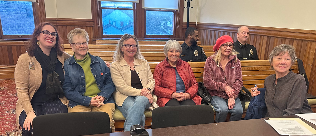 On wood benches in the City Council Chamber. 