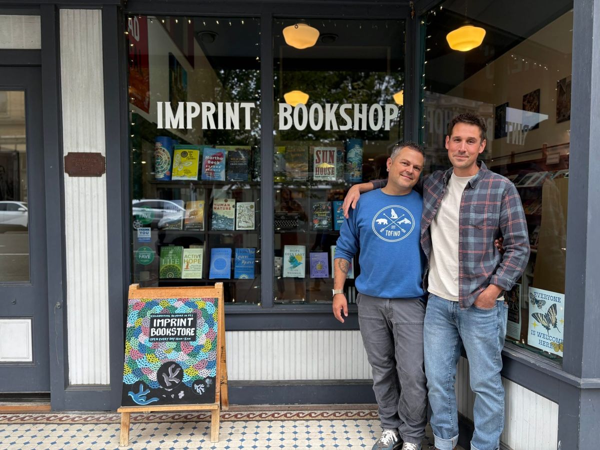 John Blomgren and Garrett Jones stand outside their Imprint Bookshop, a gray storefront window with books.