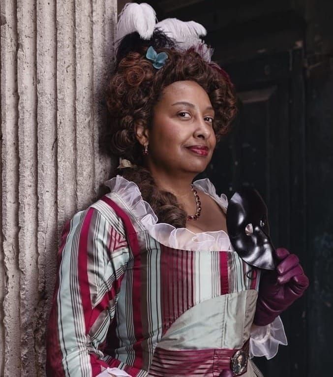 Gigi Coulson, a woman of color, in a gray and red historical dress and purple gloves, holding a mask. 