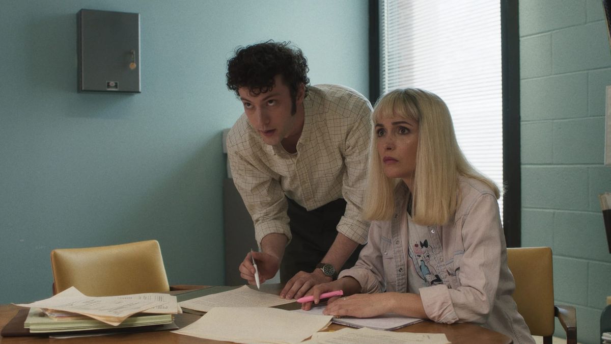 Dominic Sessa and Rose Byrne behind a desk with papers in an aqua room with tan chairs.