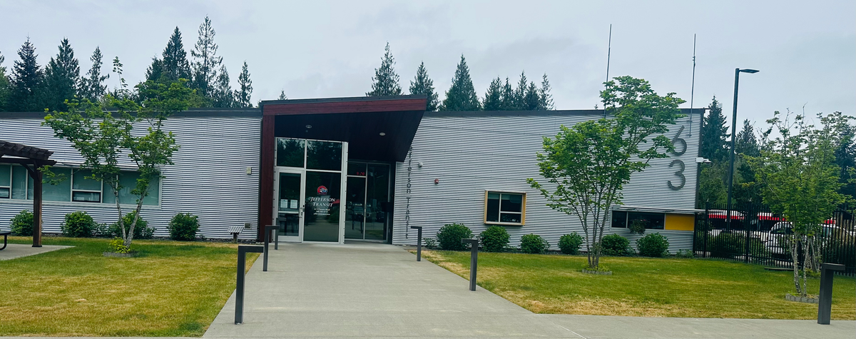A tin rhomboid building with a maroon roof-covered glass door entrance in the middle.