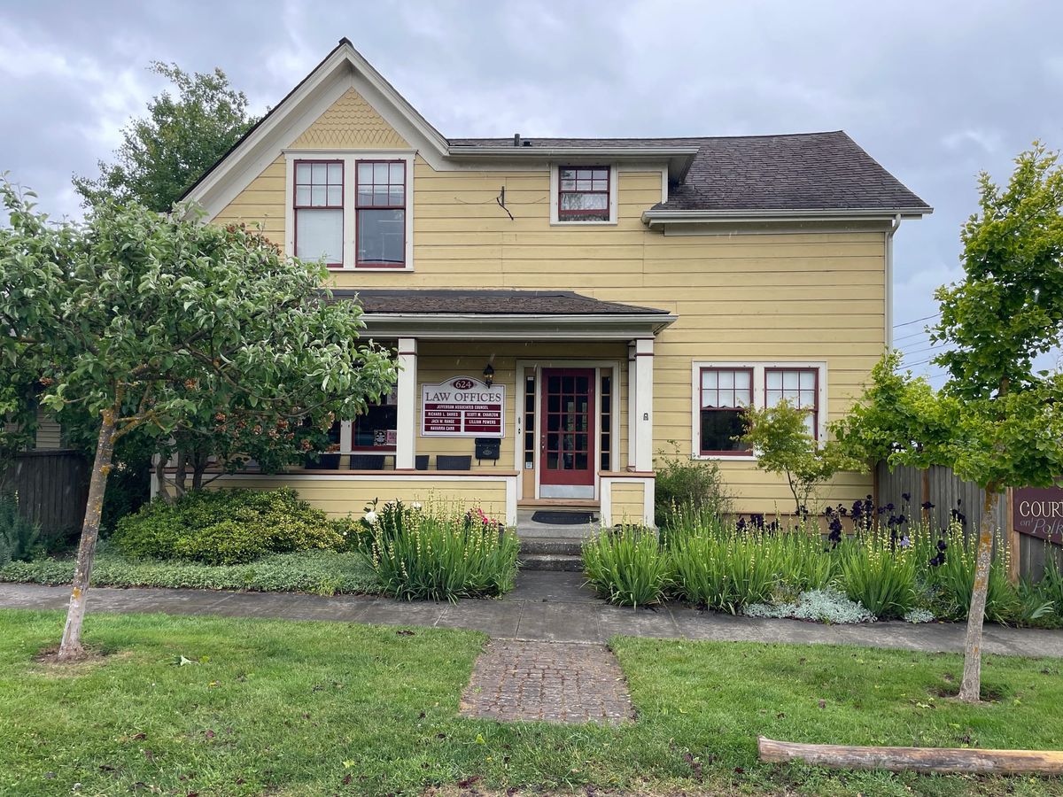 A two story yellow house with red trim converted into law offices, trees and greenery in the foreground. 