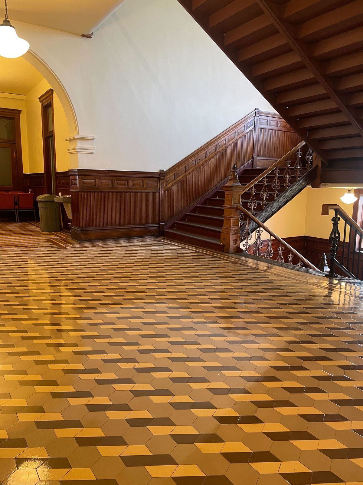 A wooden courthouse staircase, railing and wainscotting with a beige and brown patterned tile floor.