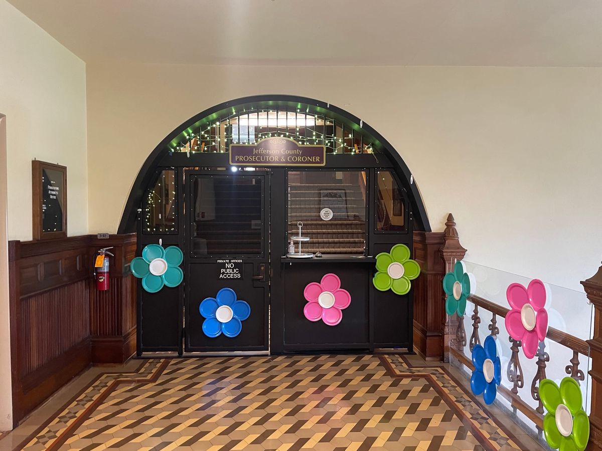 JA black painted archway with two doors. Brown and tan tile floor, and dark wood wainscotting. Colorful flowers made from paper plates are tacked everywhere.