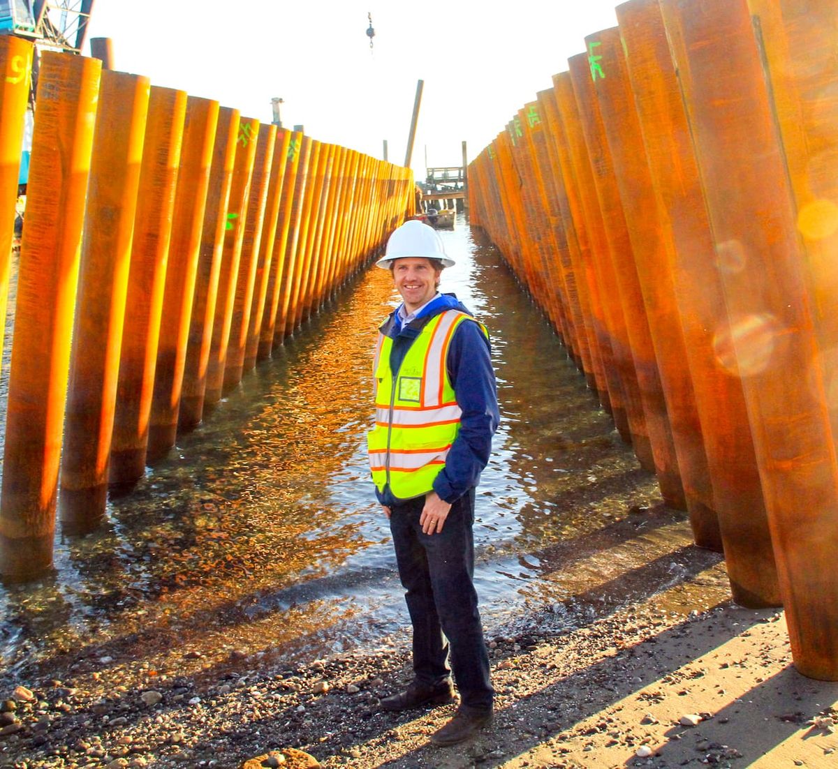 Matt Klontz, in a hard hat and reflective vest, stands in front of metal pilings on the beach of Point Hudson. 