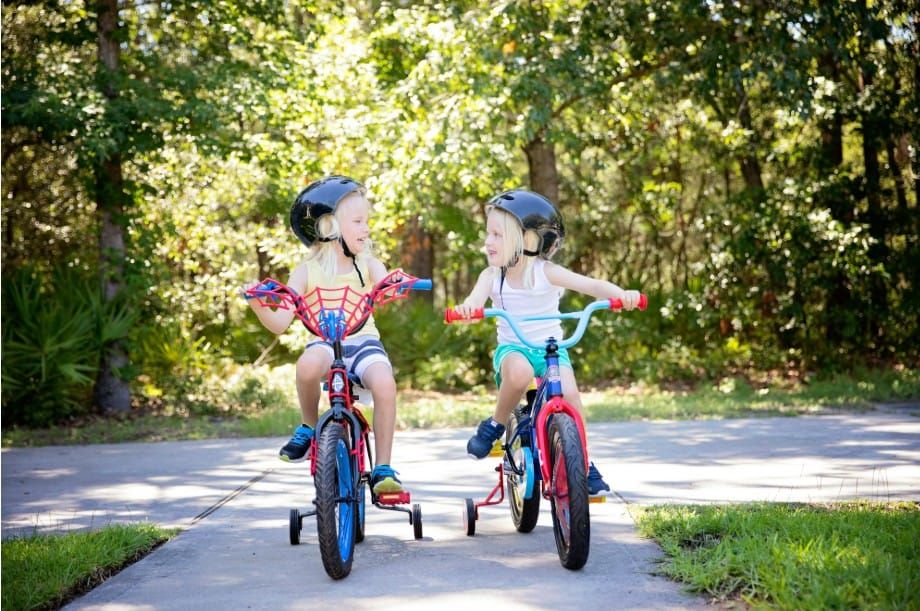 Two kids on bikes with training wheels wearing helmets at the end of a sidewalk. 