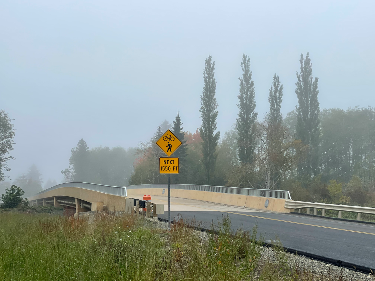 Looking north over the Twana Bridge which is tan cement with rails but no sidewalk, a yellow warning sign for pedestrians and bicyclists in the fore and green scenery in the background. 