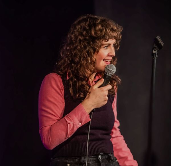 Jaci Terjeson on stage on a microphone wearing a pink shirt, maroon vest and black jeans