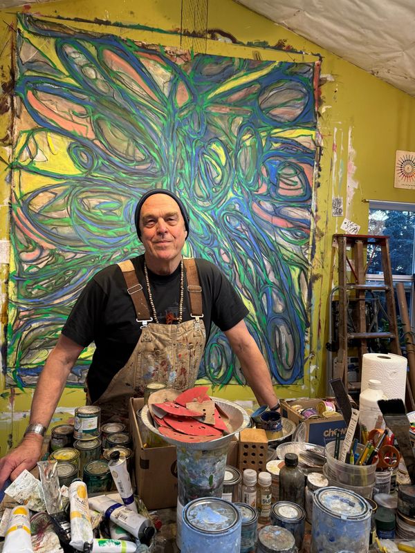A collection of paints in front of painter Randy Sturgis, who stands in front of one of his colorful paintings
