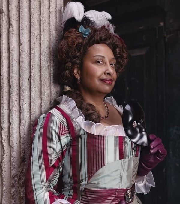 Gigi Coulson, a woman of color, in a gray and red historical dress and purple gloves, holding a mask. 