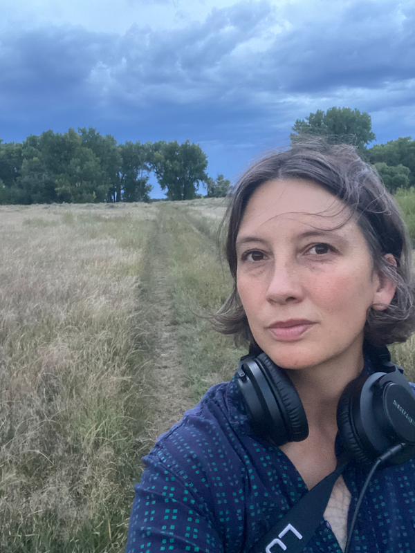 Nhatt Nichols with headphones in front of a prairie with some trees. 