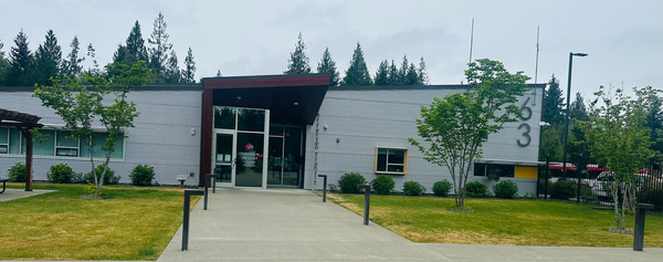 A tin rhomboid building with a maroon roof-covered glass door entrance in the middle.