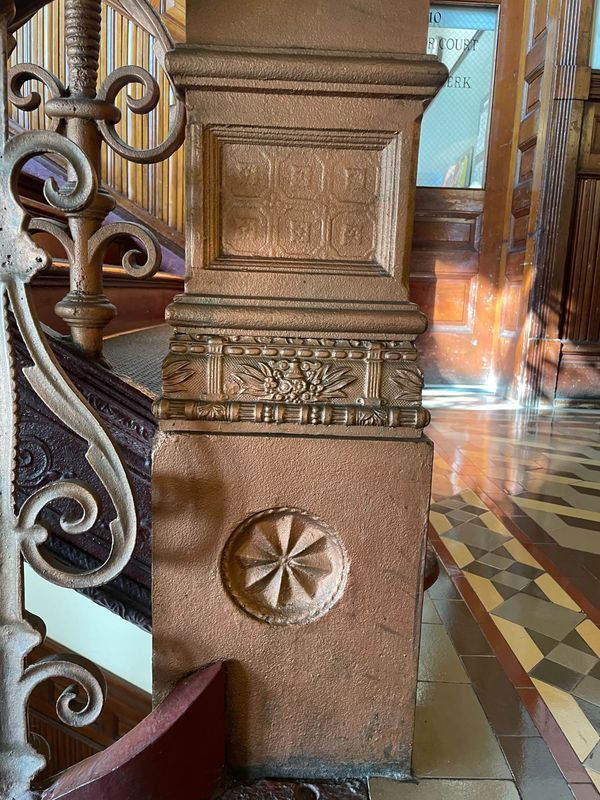 A large copper colored banister with carved details at the base of a stairway. 