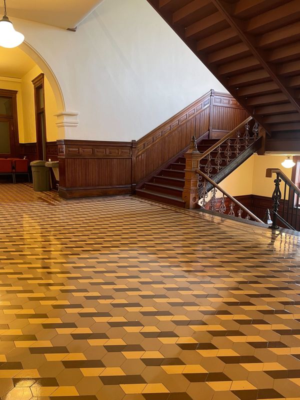 A wooden courthouse staircase, railing and wainscotting with a beige and brown patterned tile floor.