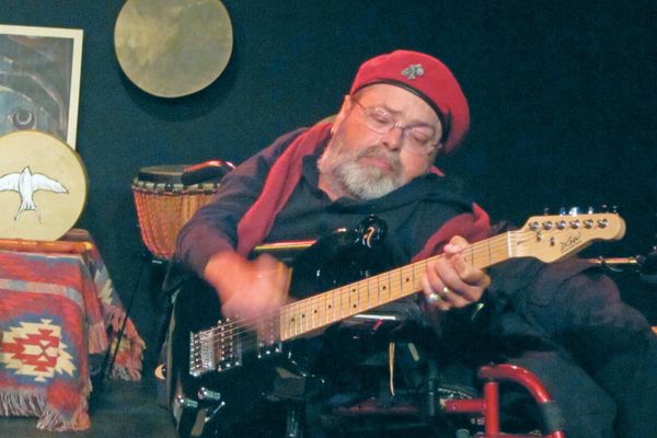 Daniel '3D' Deardorff performing with his guitar in a red beret with some drums in the background. 
