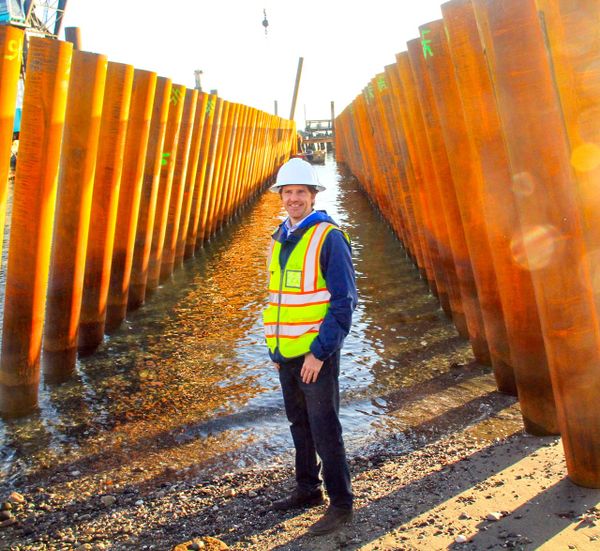 Matt Klontz, in a hard hat and reflective vest, stands in front of metal pilings on the beach of Point Hudson. 