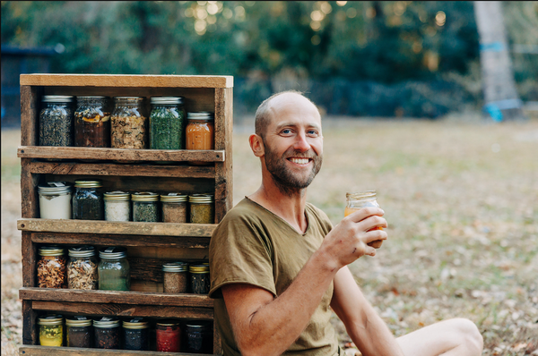 Robin Greenfield smiling and holding up a jar leaning against a shelf with lidded glass jars of food. 