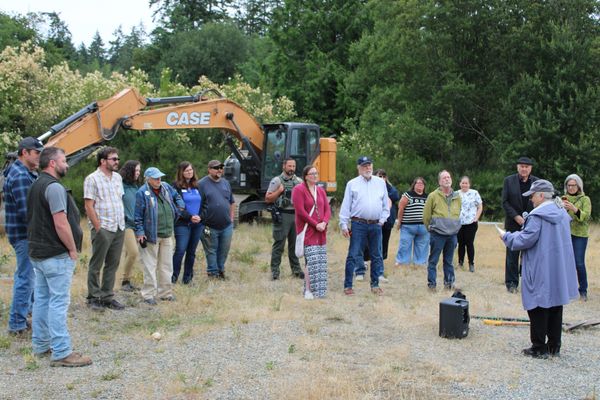 Groundbreaking ceremony on July 1, attendees looking on during a speech with an excavating tractor in the background.