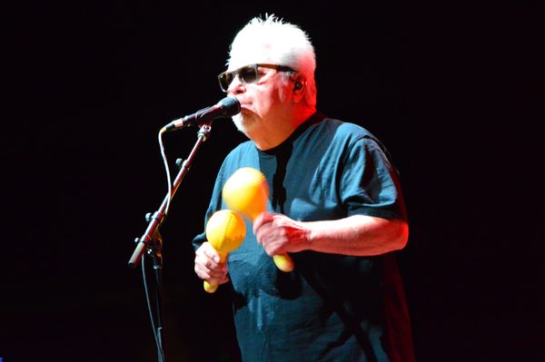Cesar Rosas singing into a microphone and shaking his maracas. 