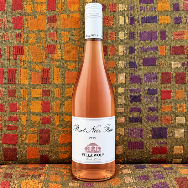 A clear bottle of Villa Wolf 2025 Pinot Noir Rosé in front of a colorfully patterned fabric background. 