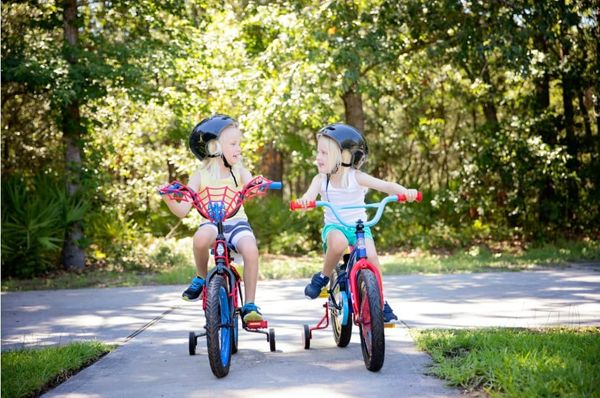 Two kids on bikes with training wheels wearing helmets at the end of a sidewalk. 