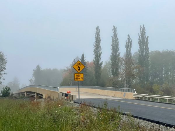  Looking north over the Twana Bridge which is tan cement with rails but no sidewalk, a yellow warning sign for pedestrians and bicyclists in the fore and green scenery in the background. 