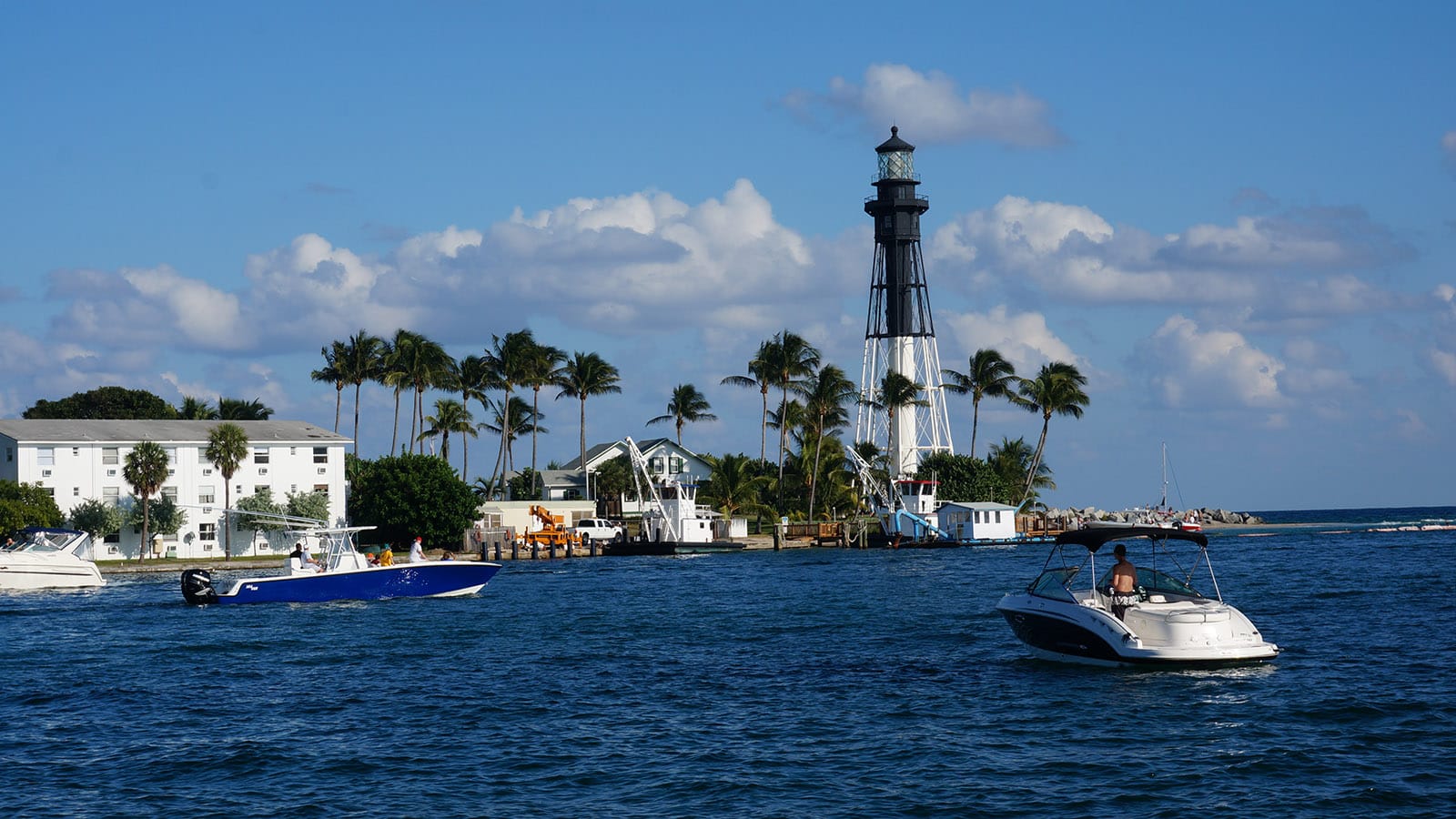 Boater in a busy channel in front of a half black half white lighthouse at an inlet