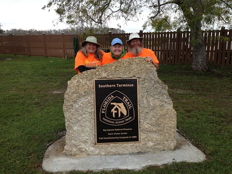 Two women and a man behind a large stone with the Florida National Scenic Trail Southern Terminus plaque