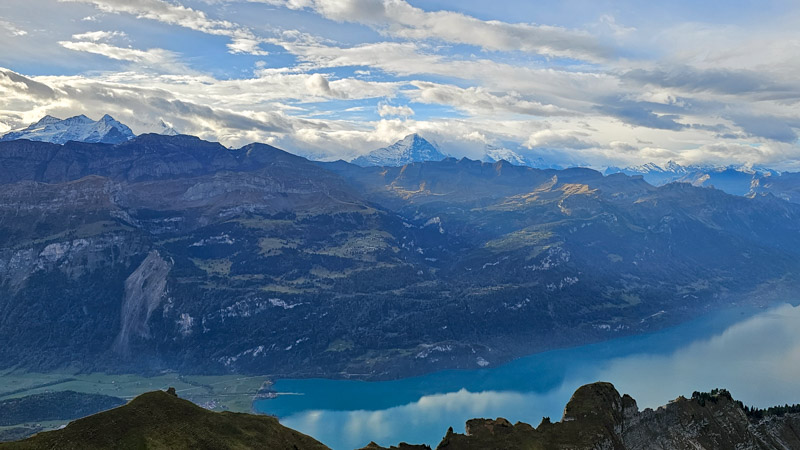 Panoramic landscape view of the Swiss Alps, featuring snow-capped mountain peaks in the distance, rolling hills covered with forests and patches of greenery, a large turquoise lake, and a cloudy blue sky.