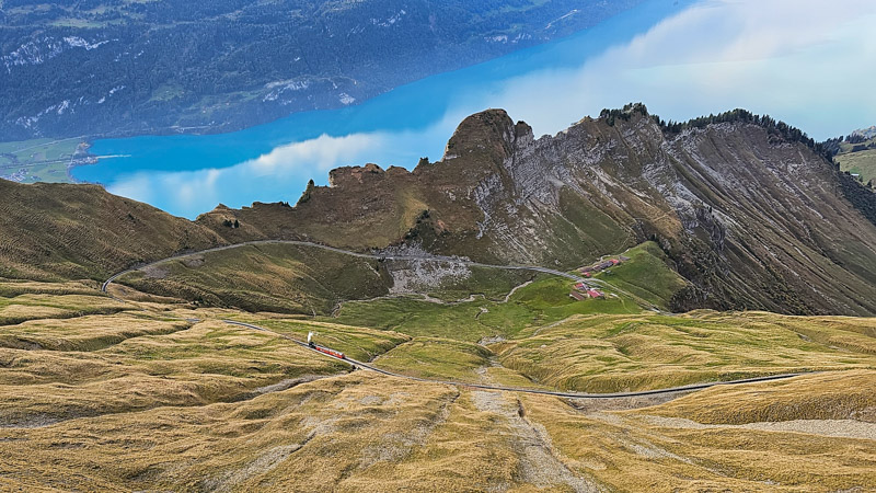 Aerial view of a mountain landscape featuring a red train winding along a curved track amidst green and brown grassy slopes, with a large turquoise lake and distant mountains in the background.