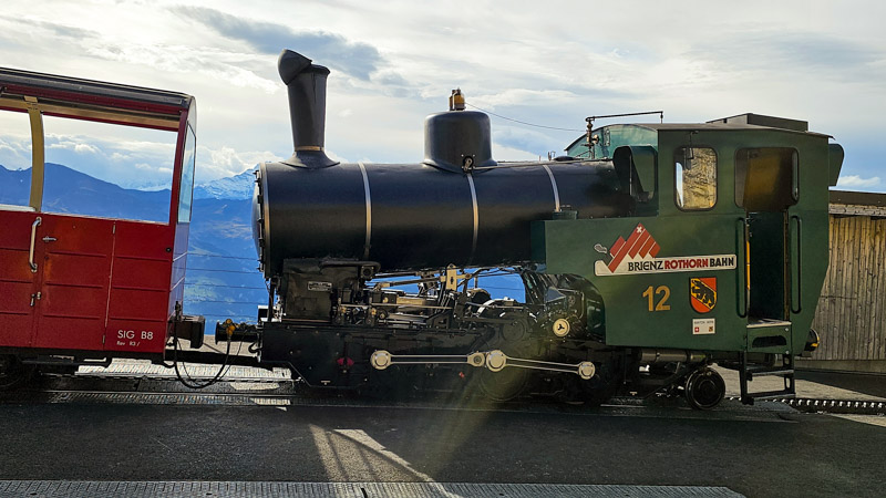 Black steam train with green cab and the words "Brienz Rothorn Bahn" and number "12" on its side, coupled to a red passenger car, with a mountainous landscape in the background.