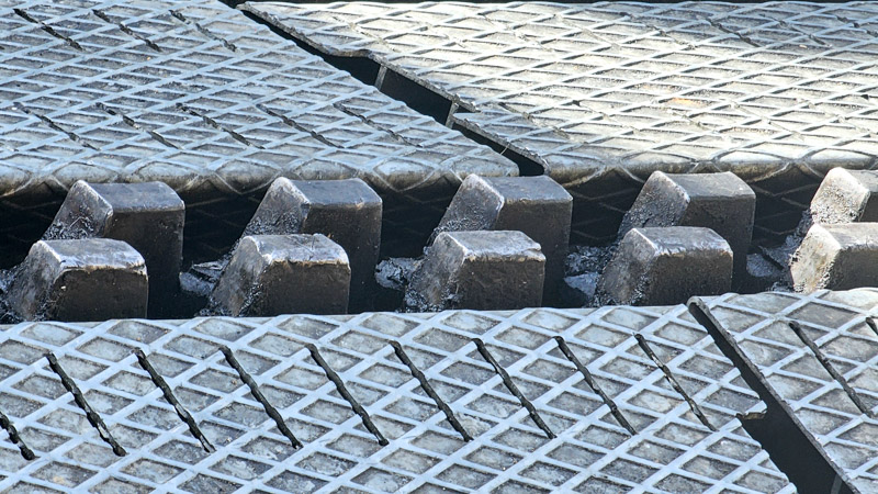 Close-up of industrial metal grating with diamond-patterned surface, interspersed with evenly spaced, irregularly shaped dark grey cog teeth.