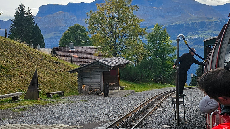 A man in black standing on a small platform connected to a pole, refilling a steam locomotive with water from a hose. The train, on tracks, is in a mountain setting, with wooden structures and a mountain in the background.
