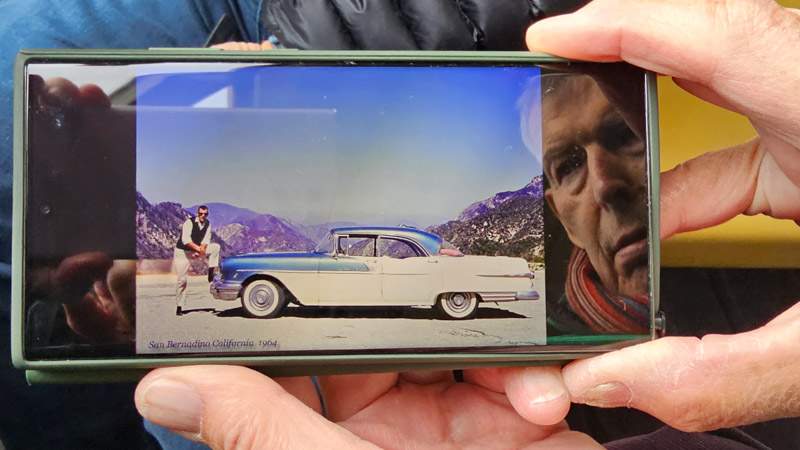 Person holding a phone displaying a vintage photograph from 1964 of a man leaning on a blue and white classic car in San Bernardino, California, with mountains in the background; the phone's screen also reflects the holder's face.