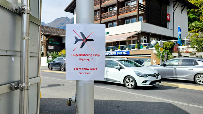 A sign on a light pole in a Swiss town announces that the Axalp flight show is canceled; the sign depicts a black airplane symbol with a red slash through it. Cars and buildings line the street in the background.