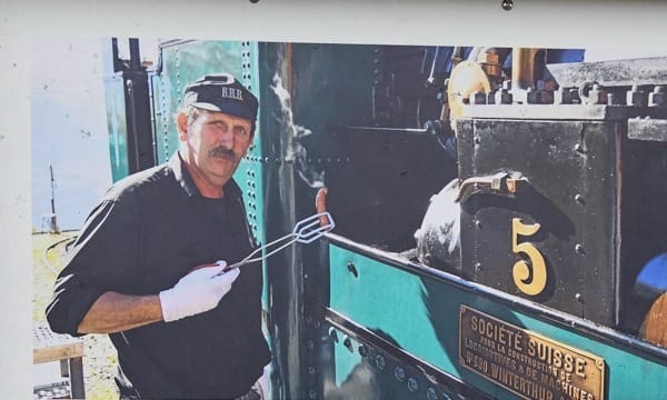 Man in cap and gloves holding tongs with a steaming sausage firmly grasped in front of a vintage steam train, with the number 5 visible.