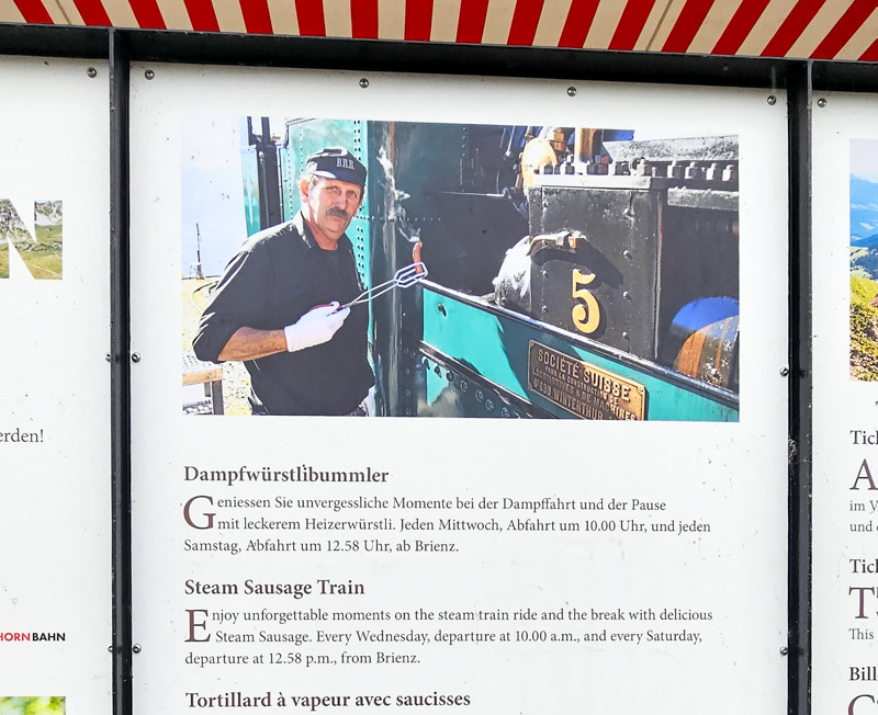 A man wearing a "BRR" hat, white gloves, and a black shirt holds grilling tongs over a small, vintage, green and black steam train labeled "Société Suisse" with the number "5" on the side, advertising "Steam Sausage Train" in both German and English.