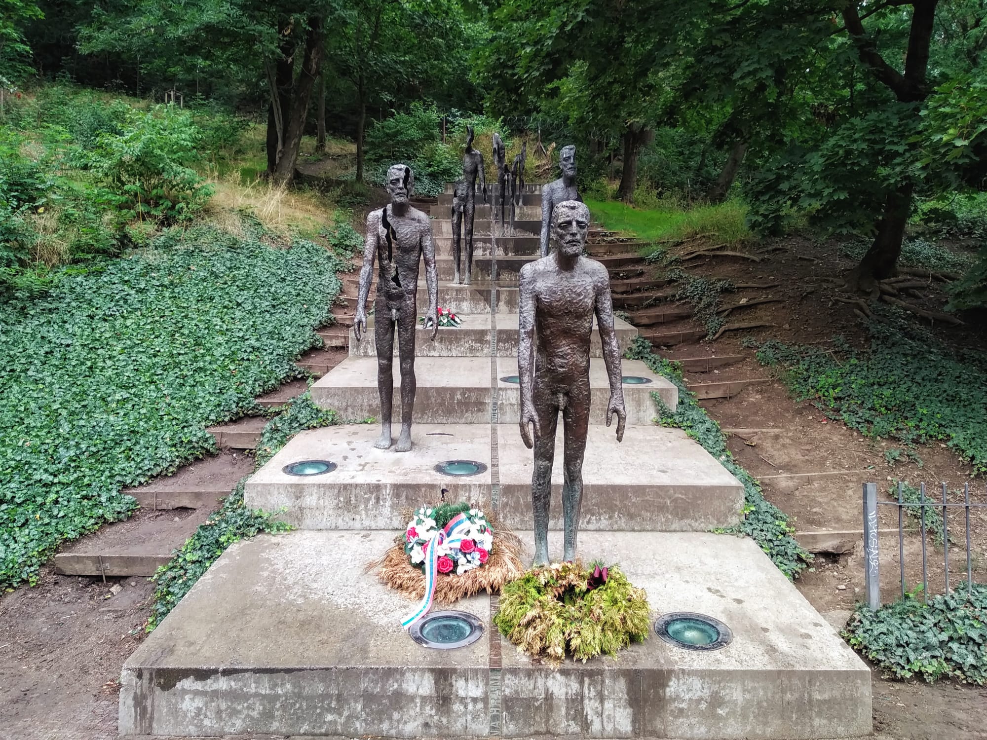 Bronze statues of gaunt human figures on concrete steps, part of a memorial in a wooded area; each figure stands stark naked with visible ribs and pained expressions, some hold flowers, while wreaths lay at the base of the steps.