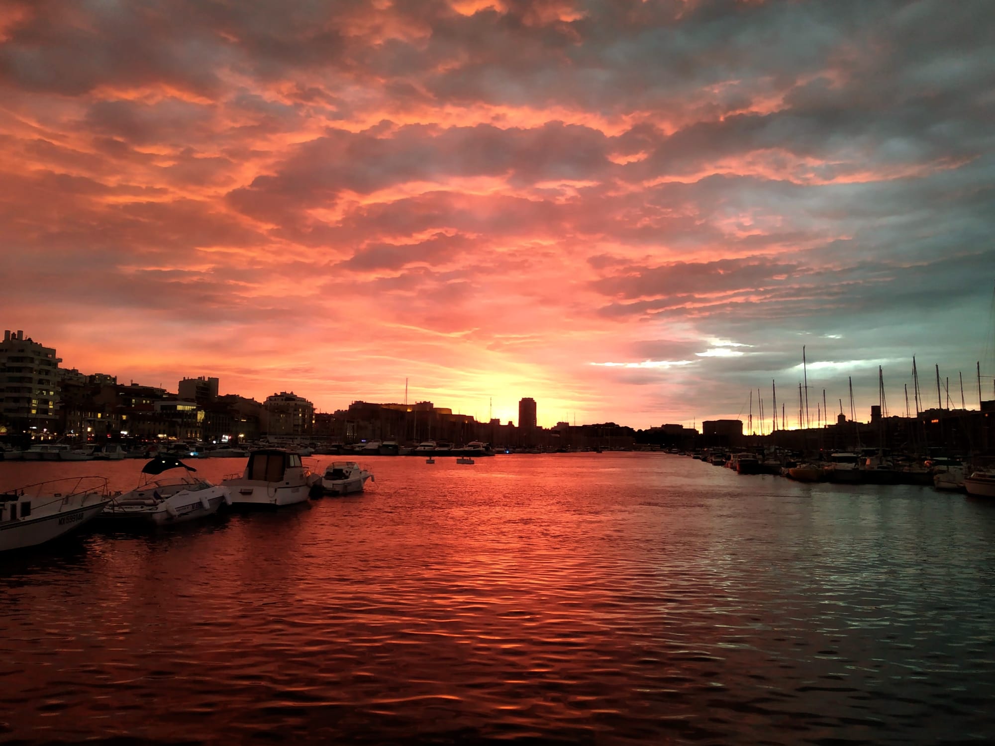 Vibrant sunset over a harbor with boats and buildings silhouetted against a sky of fiery orange and pink clouds reflecting in the water.