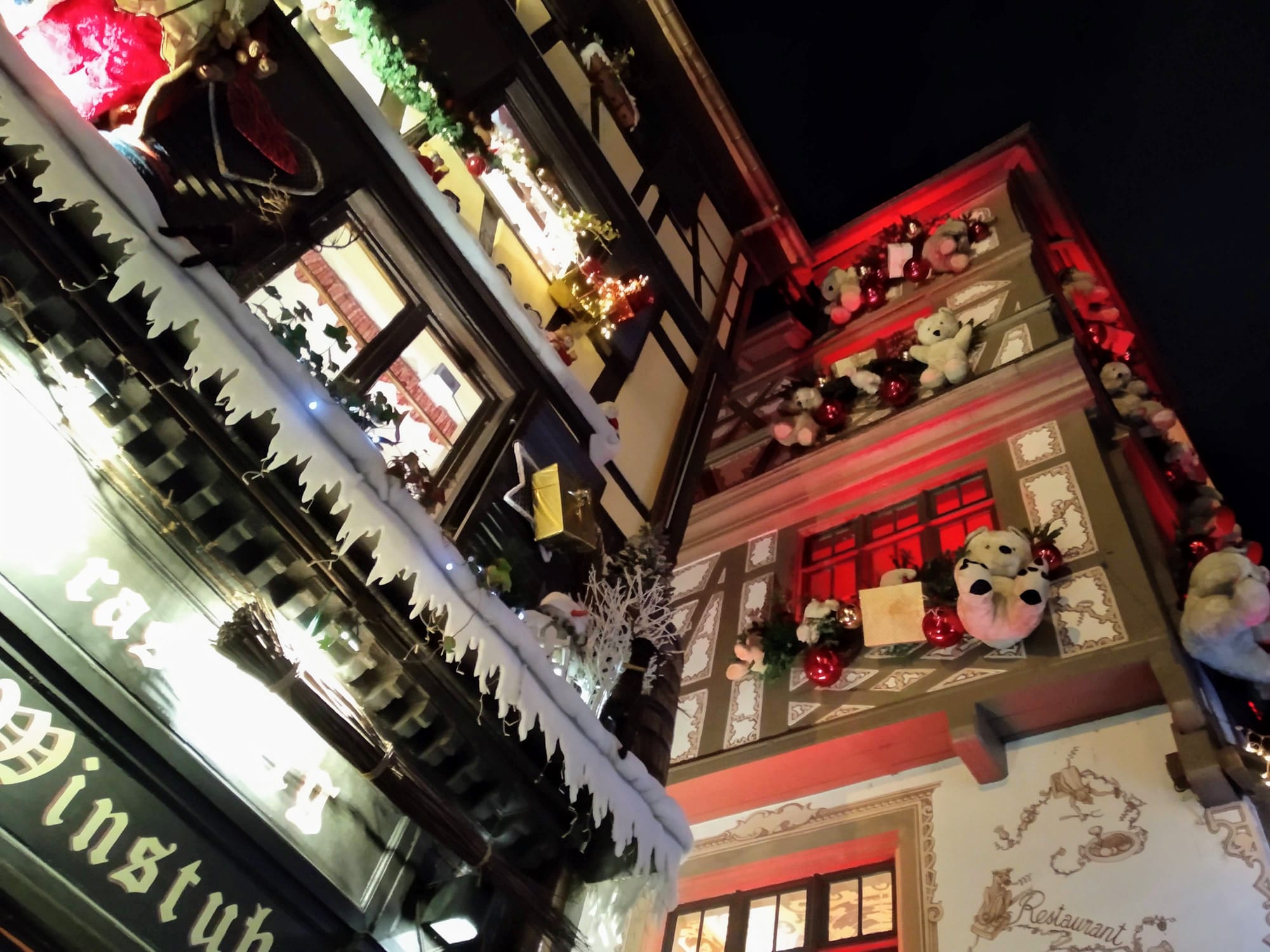 Ornate half-timbered buildings at night, decorated with Christmas lights, garlands, and teddy bears. Icy white trim and "Winstub" signage visible.