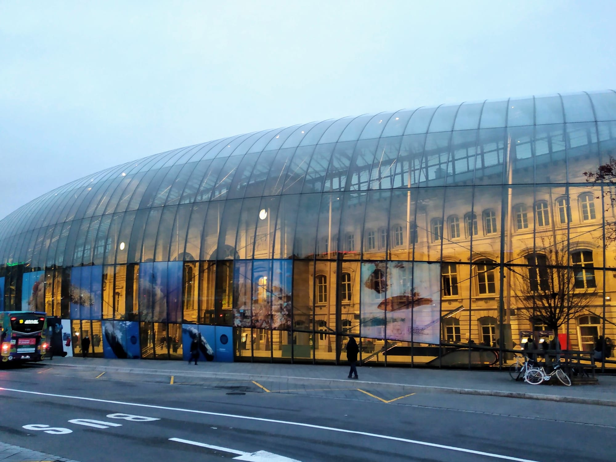The beige-coloured Gare de Strasbourg and its arched windows behind a curved, glass roof. Large panels displaying underwater images line the lower section of the glass exterior under a cloudy morning sky.