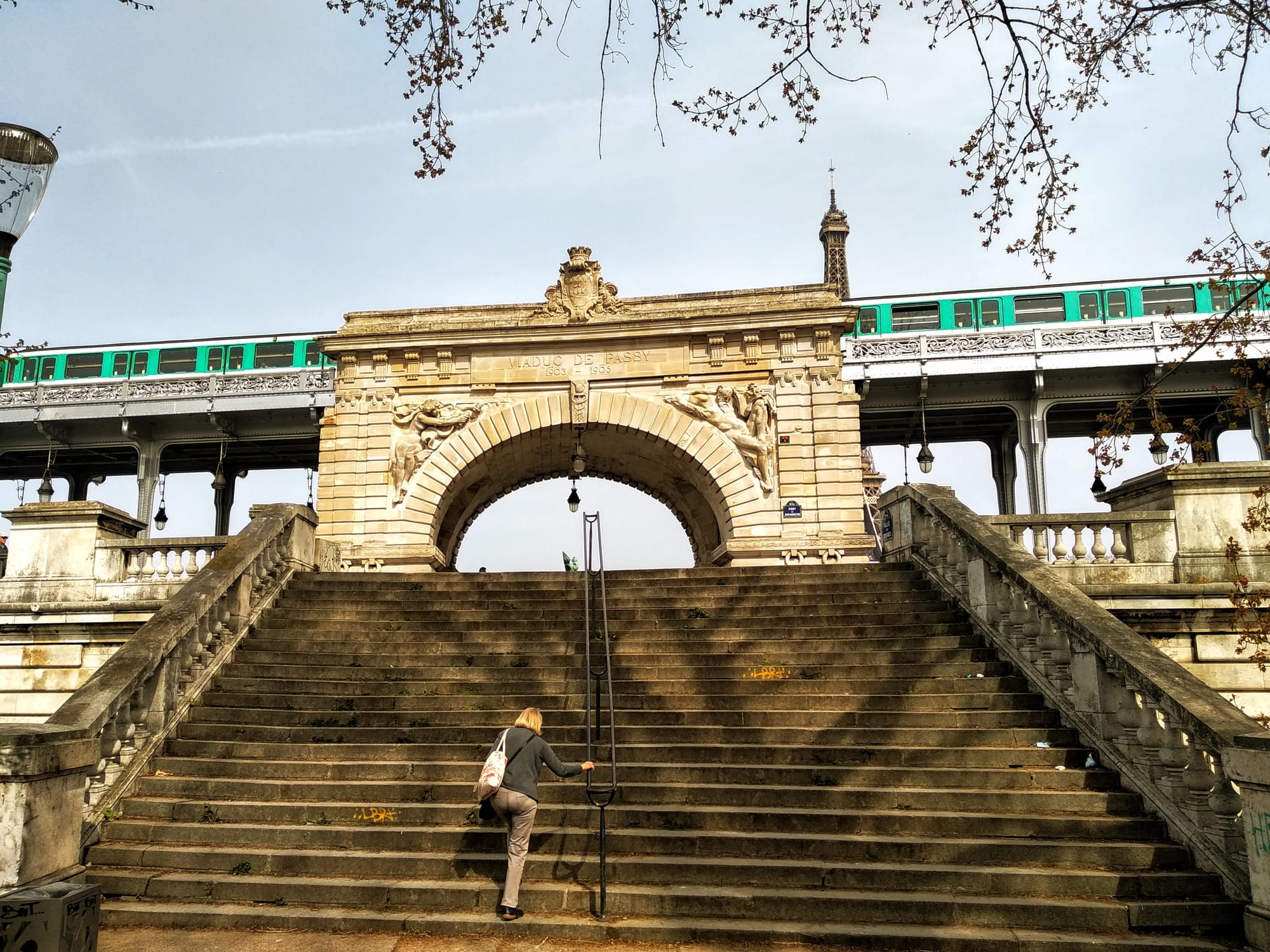 Woman ascending the monumental stone staircase of the Bir-Hakeim bridge in Paris, with the elevated metro train passing overhead, the bridge's archway, and the Eiffel Tower visible in the background.