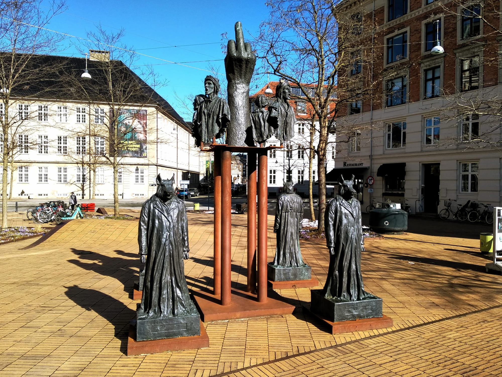 On the ground stand four life-size bronze versions of Galschiøt's famous sculpture The Inner Pigdog, and above them float four busts of Dante himself. In the middle of this square construction stands a Greek column with a very large fuck finger on top.