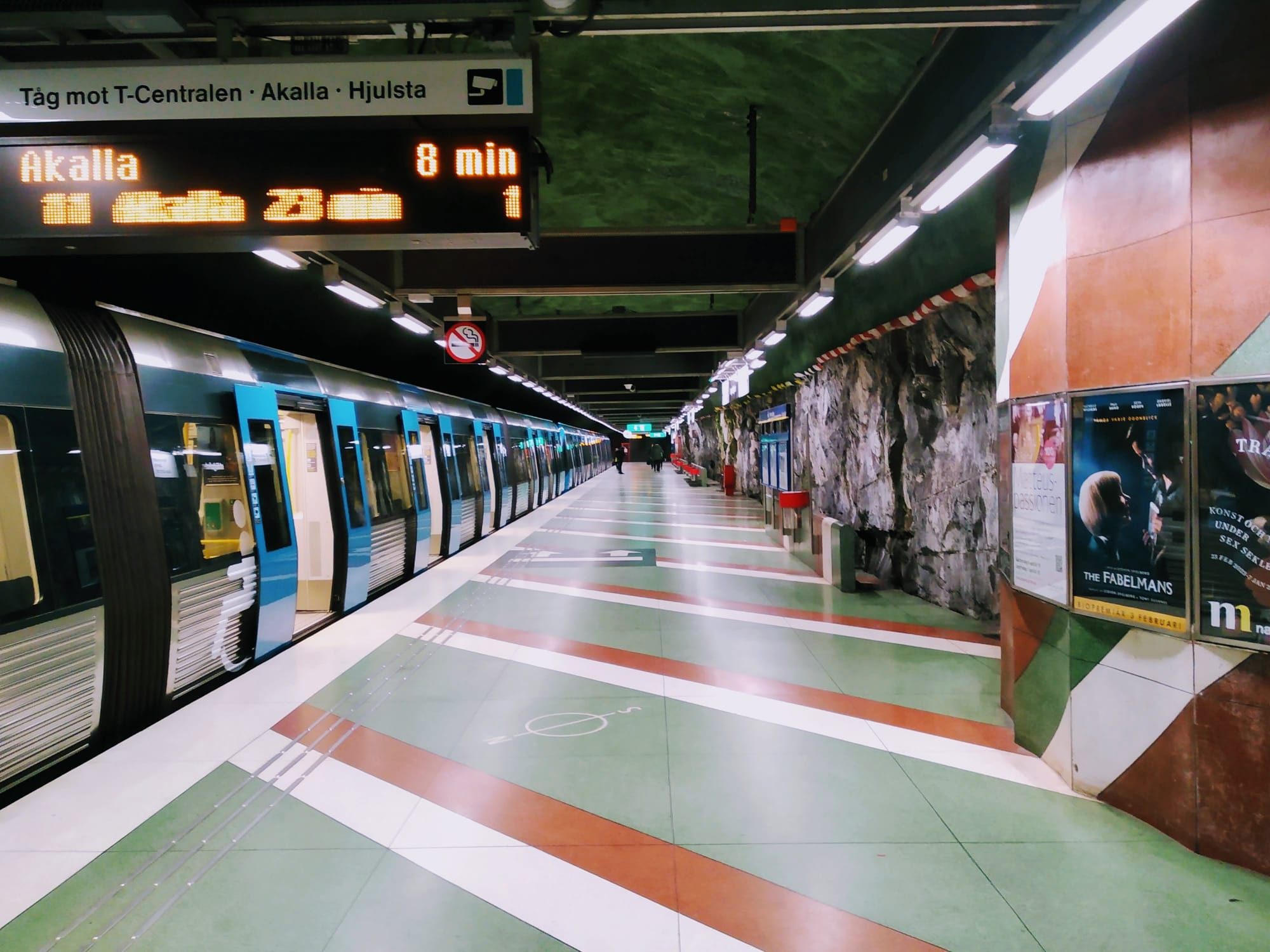 An underground train to Akalla waits for passengers alongside red, white and green patterns on the platform.